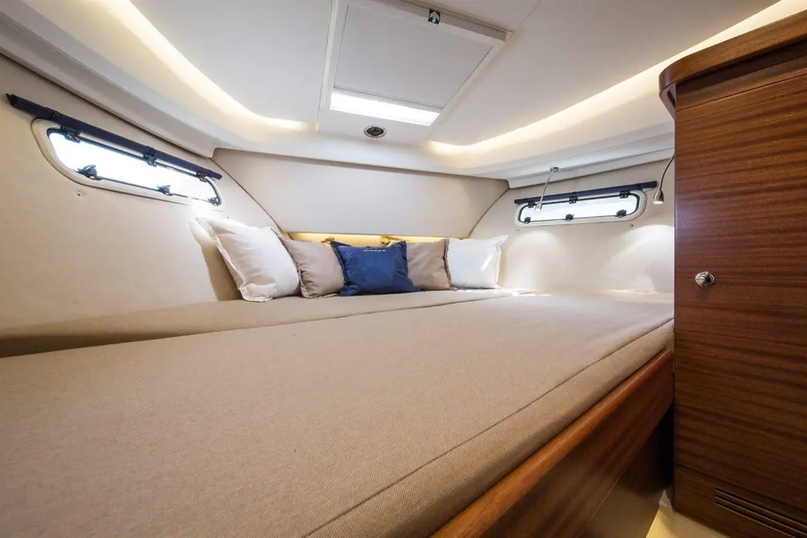 A modern yacht cabin features a large cushioned bed with decorative pillows, natural light from porthole windows, and wooden cabinetry.